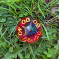 Top down view of a percentile die with a blue center, red exterior. and gold numbers, sitting in a bed of clover, grass, and dandelion greens